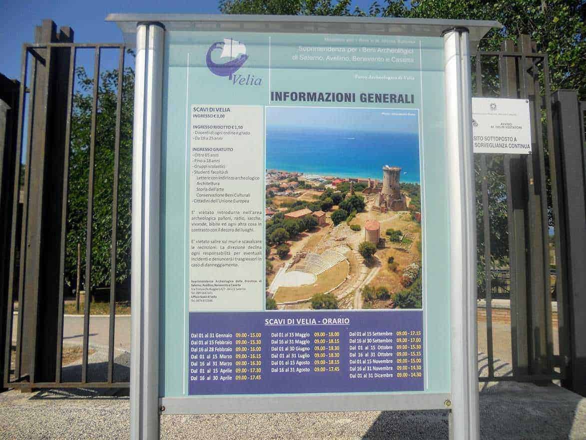 Pannello informativo esterno con panoramica archeologica di Velia, indicando orari di apertura e principali attrazioni, immerso in un ambiente naturale.