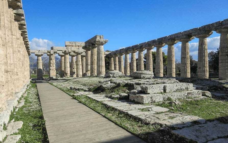 Antichi resti di templi greci con colonne in stile dorico, sito archeologico in perfette condizioni all’aperto, con un sentiero pedonale e un cielo azzurro sullo sfondo.