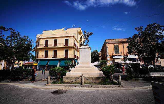 FOTO: Monumento storico e piazza centrale a Civitavecchia, Italia, con edifici ottocenteschi e statua in centro, situazione urbana vivace, zona turistica e commerciale.