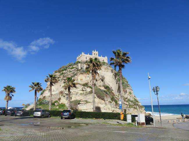Tropea Borgo dei borghi 2021 in Calabria! 8 Vista panoramica di un castello sulla scogliera con palme sulla costa, in una giornata soleggiata con cielo blu e mare azzurro, ideale per fotografie turistiche e attrazioni storiche in Italia.