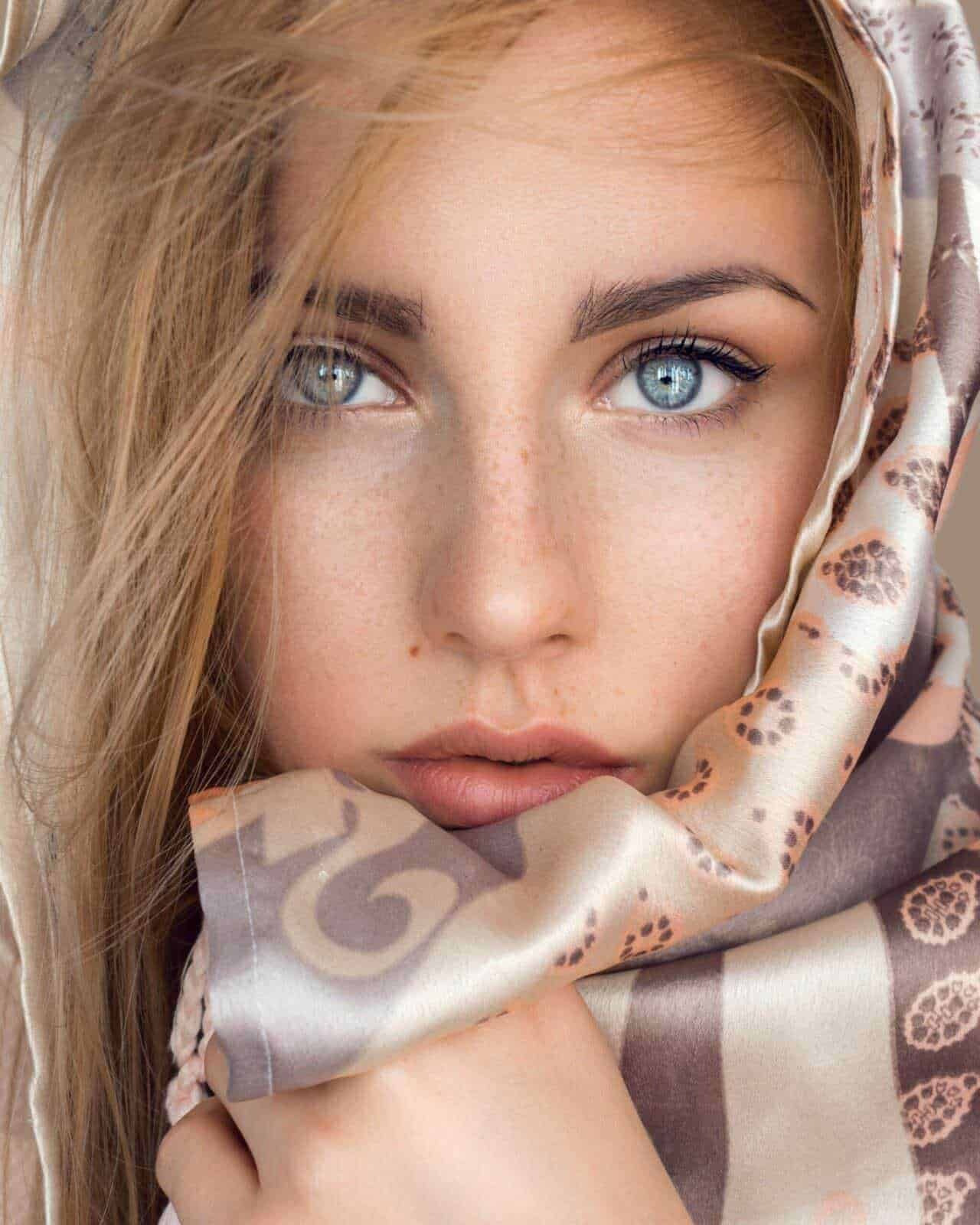 Affascinante volto di donna con occhi azzurri e pelle chiara, avvolta da un foulard floreale in raso, stile glamour e moda. Perfetto per blog di bellezza e moda, con focus su skincare e tendenze di stile.