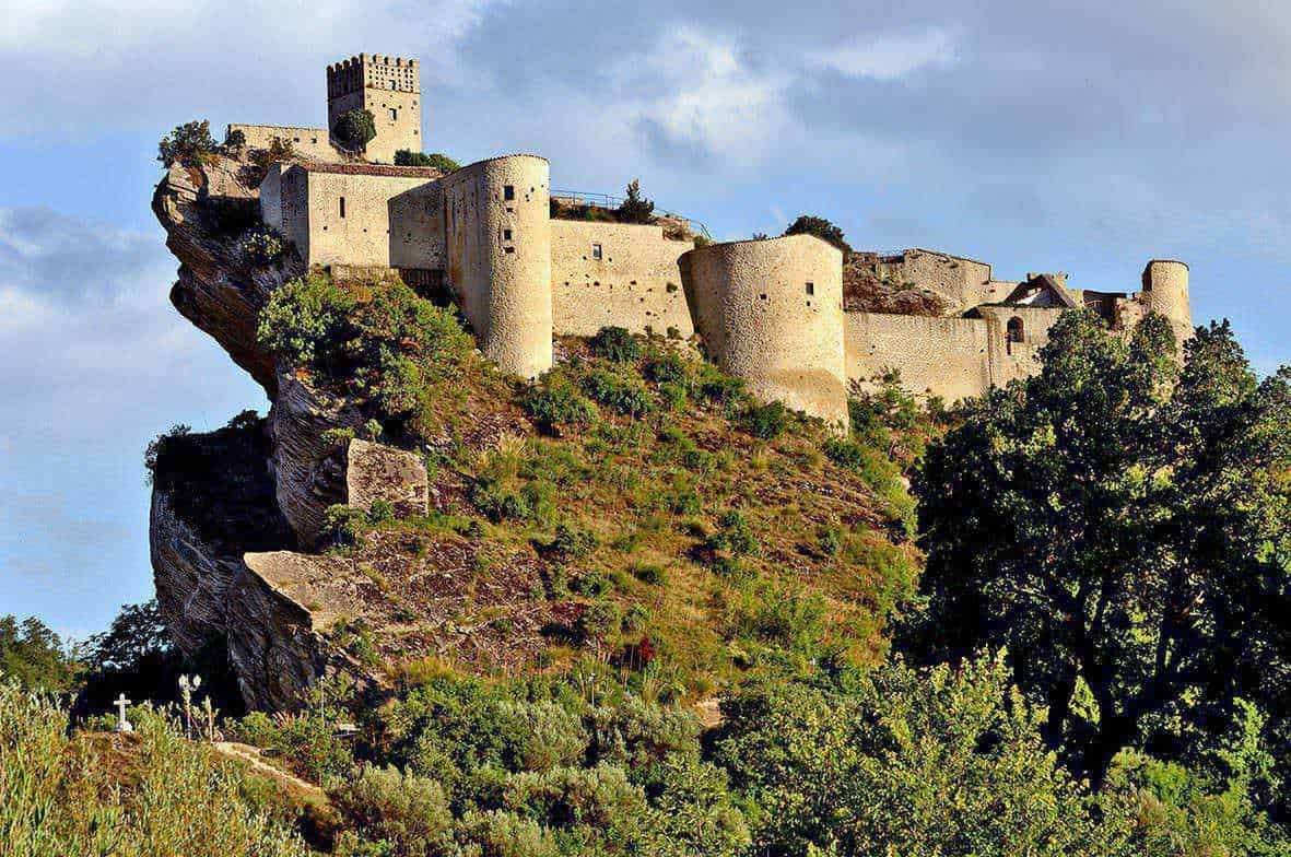 Castello medievale su scogliera con vegetazione circostante e cielo nuvoloso, esempio di architettura storica italiana e attrazione turistica.