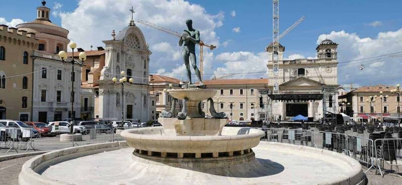 Dove andare in Abruzzo montagna estate 8 Fonte Piazza con fontana e statue al centro, scena urbana di Cagliari con edifici storici e palco per eventi, cielo sereno e traffico, rappresentativa del cuore della città.