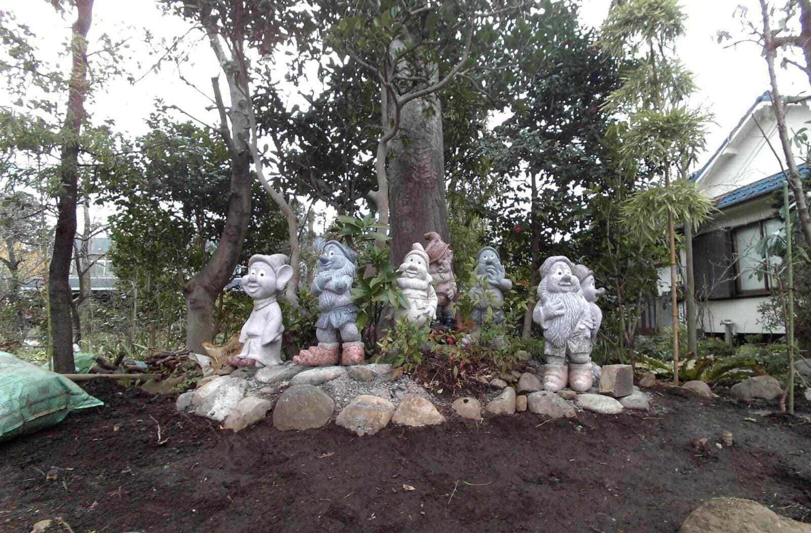 Colorata statua di sette nani della fiaba di Biancaneve in giardino, circondata da piante e alberi, con una casa sullo sfondo, creando un’atmosfera fiabesca e naturale.