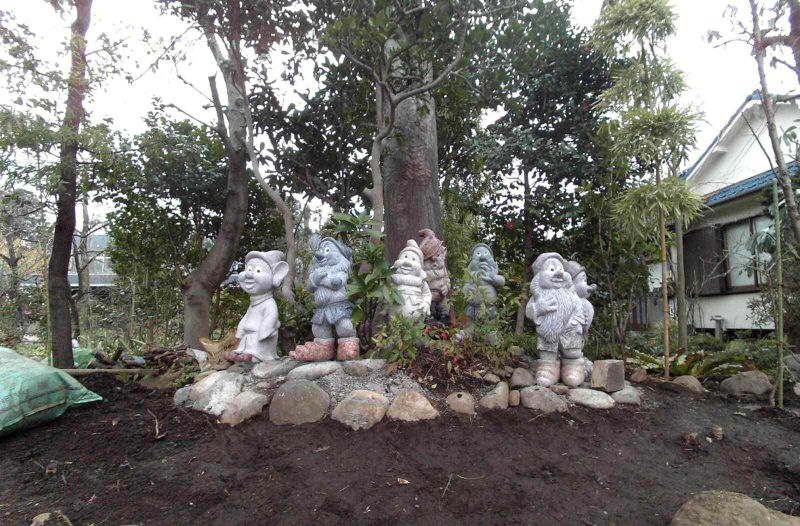 Origine Nani da giardino da antica Roma a oggi! 4 Colorata statua di sette nani della fiaba di Biancaneve in giardino, circondata da piante e alberi, con una casa sullo sfondo, creando un’atmosfera fiabesca e naturale.