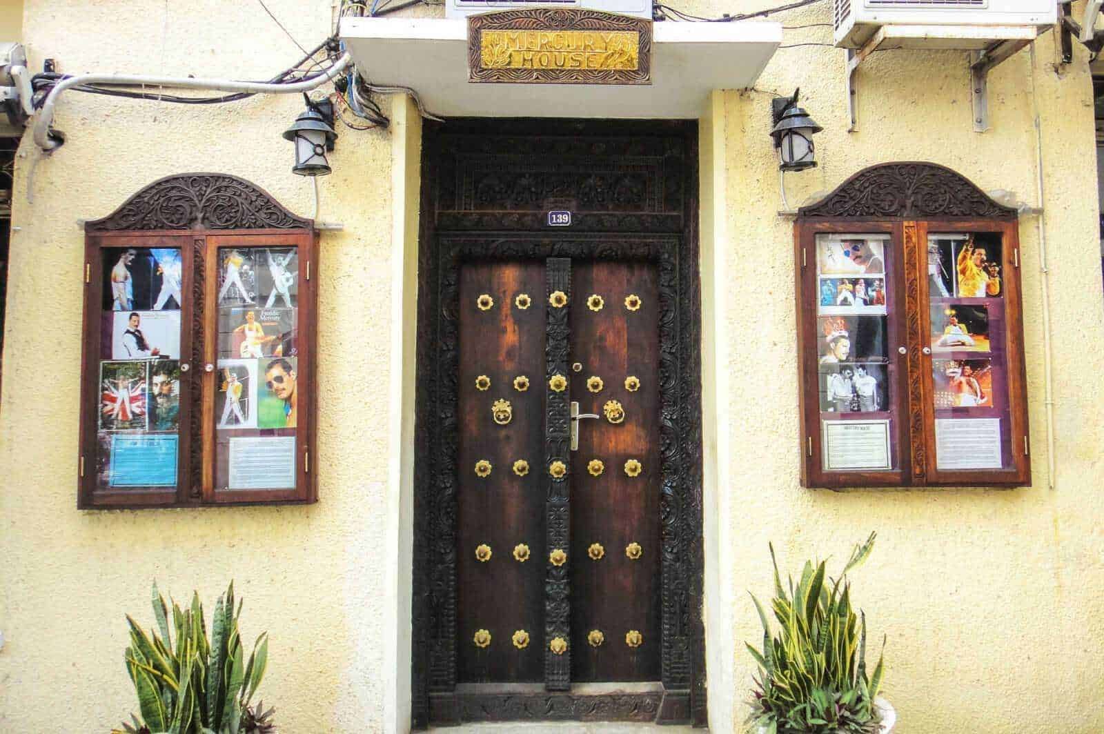 Porta d'ingresso decorata con dettagli in ferro battuto e decorazioni floreali, inserita in un edificio con pareti di colore giallo e vetrine decorative con foto di celebrità e manifesti culturali.