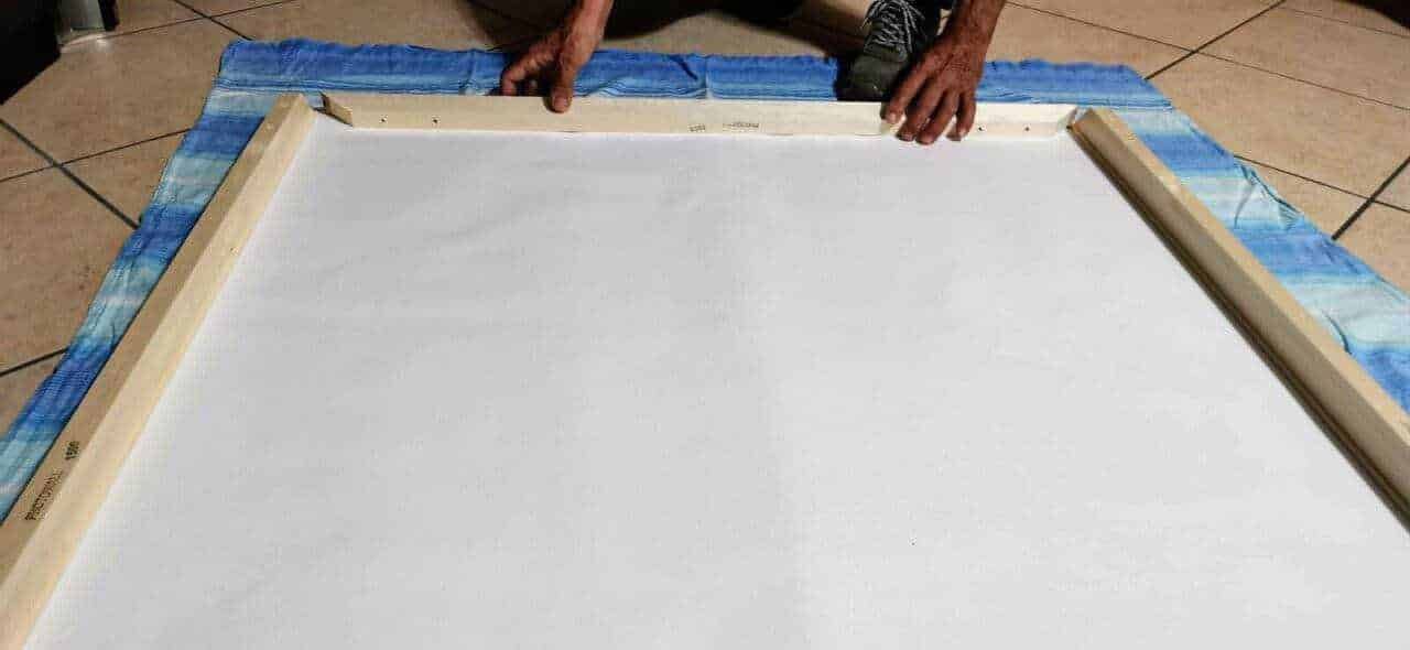 Tela di supporto per dipingere o incorniciare con cornice di legno, posizionata su un telo di plastica blu su un pavimento in piastrelle. Strumento di bricolage in uso per lavori artistici o decorativi.