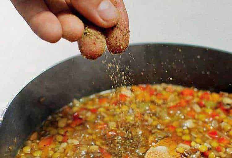 Soffritto in padella con spezie e aromi, con mano che aggiunge pepe nero in polvere, durante la preparazione di un piatto di cucina casalinga italiana.