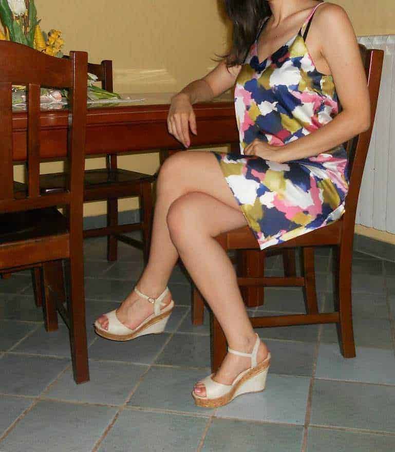 Sandali con zeppa per donna in un ambiente domestico, seduta su una sedia di legno con un vestito estivo colorato, scena di vita quotidiana e moda femminile.