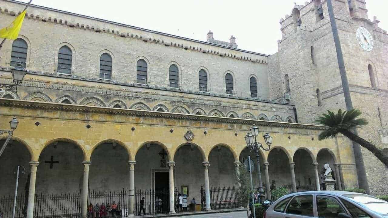 Antico edificio storico con portico e torre con orologio, situato in una suggestiva piazza italiana, con palme e lampioni, zona di grande interesse culturale e turistico, rappresenta un esempio di architettura medievale.