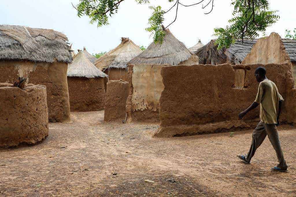 Paesaggi agricoli tropicali e subtropicali: Vecchi alloggi tradizionali in terra e paglia nel villaggio africano, con capanne dal tetto di paglia e strutture di argilla, immerso nella natura, con un uomo che attraversa la strada sterrata.