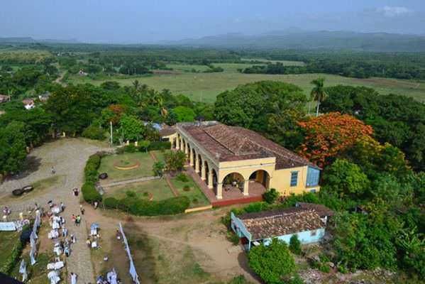 Vecchio edificio coloniale in mezzo alla natura lussureggiante, circondato da alberi e campi verdi, con persone in attività all'aperto e un'ampia area aperta.