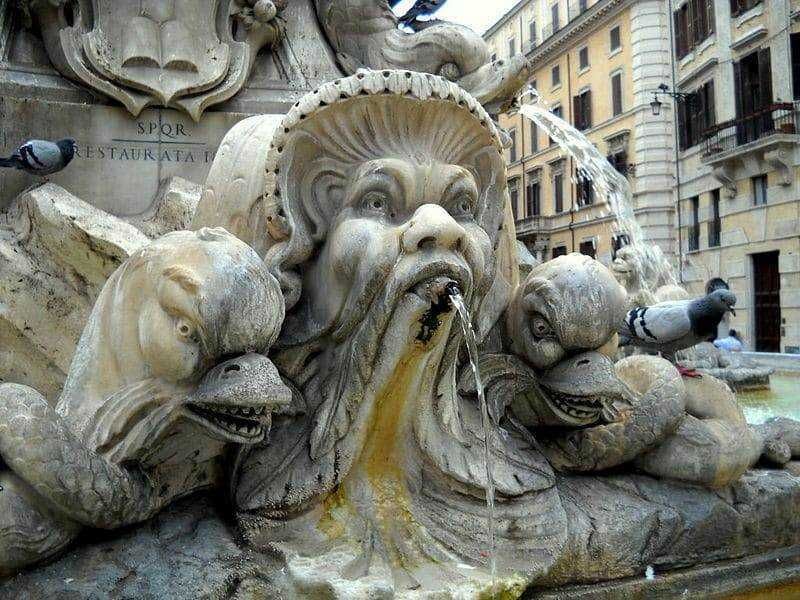 Antica fontana in Piazza Navona a Roma con sculture di teste di figure mitologiche e animali marini, sfondo con edifici storici e architettura barocca, simbolo del patrimonio artistico italiano.