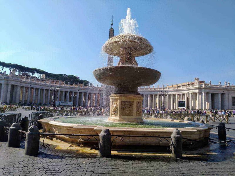13 Fontane storiche di Roma celebrative 14 Fontana di Piazza San Pietro a Vatican City, simbolo di Roma, con statua decorata e acqua che spruzza verso il cielo, circondata da edifici storici e visitatori, esempio di arte e architettura classica.