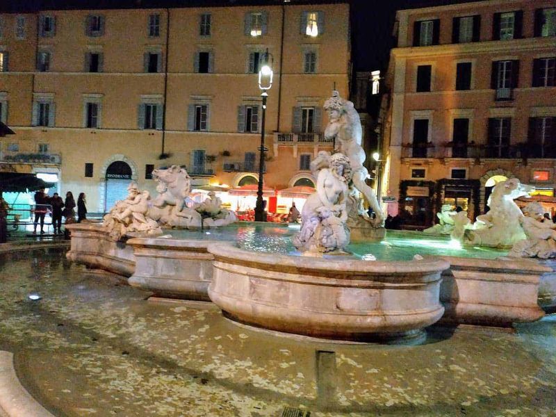 13 Fontane storiche di Roma celebrative 8 Fountain with statues in Piazza Navona, Rome at night illuminated by streetlights, lively atmosphere with tourists and locals, baroque architecture background.