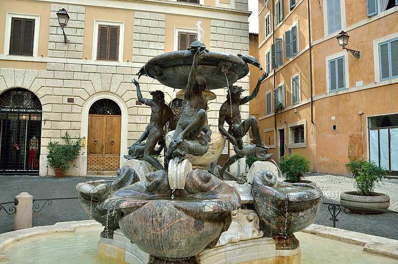13 Fontane storiche di Roma celebrative 9 Fontana artistica con sculture di figure umane su piazza antica in centro città, edifici storici e piante ornamentali sullo sfondo, esempio di arte pubblica italiana.