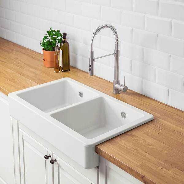 Lavandino da cucina doppio in ceramica con rubinetto professionale, posizionato su un piano in legno con sfondo in piastrelle bianche, ideale per ambienti moderni e funzionali.