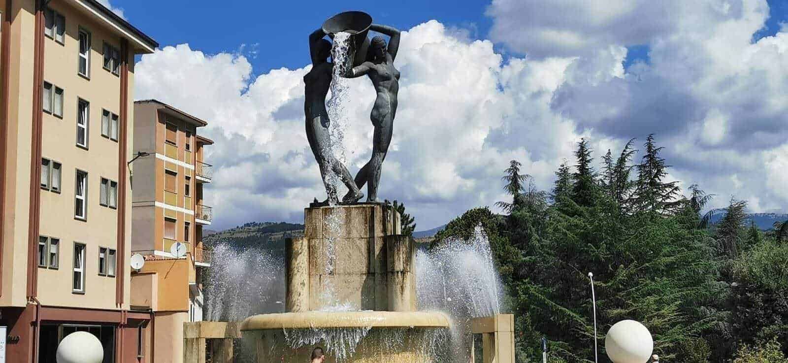 Fonte con statua di sculture femminili con acqua, nel centro di una piazza urbana, circondata da edifici e alberi, sotto un cielo nuvoloso.