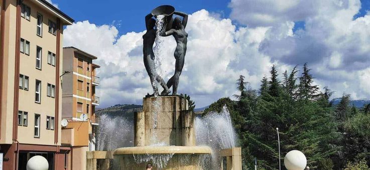 Fontane e acqua potabile: origini dall'antichità ai tempi moderni 2 Fonte con statua di sculture femminili con acqua, nel centro di una piazza urbana, circondata da edifici e alberi, sotto un cielo nuvoloso.