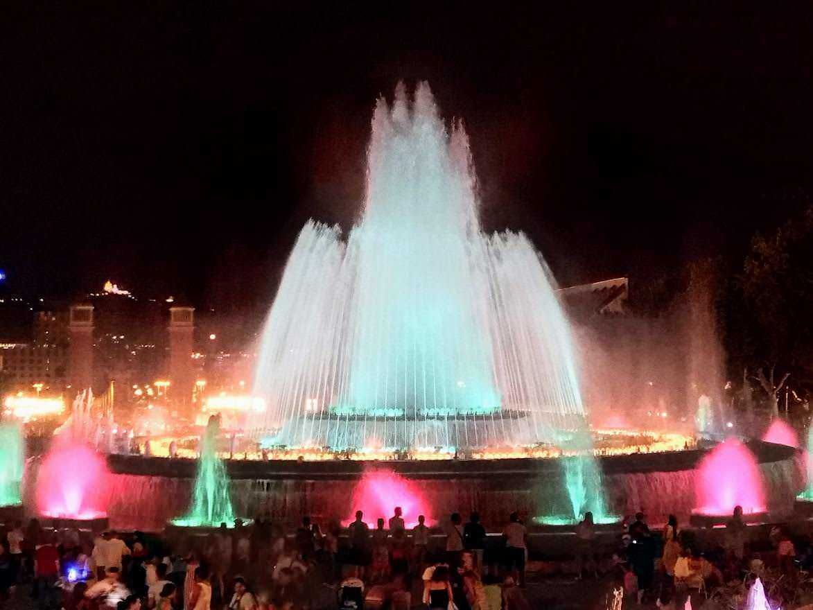 Fontana luminosa di notte con giochi d'acqua colorati e pubblico che ammira lo spettacolo, illuminata da luci multicolori, ambientata in un'area urbana di sera.