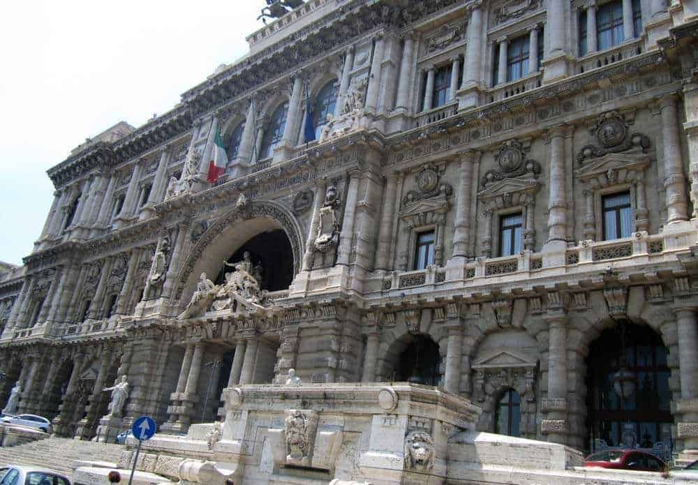Maestro di scuola superiore si trova davanti a un edificio storico riccamente decorato con sculture e colonne. La facciata si presenta imponente e dettagliata, con bandiere italiane che sventolano in alto.
