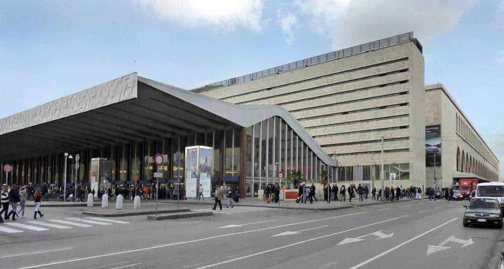 Stazione ferroviaria moderna con grande ingresso in vetro e afflusso di passeggeri, situata in un'area urbana affollata con traffico veicolare. Presenta un'architettura innovativa e funzioni di trasporto pubblico.