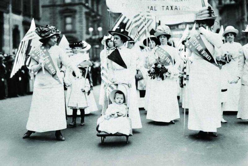 Storia del rossetto rosso nel tempo 5 Manifestazione storica femminile con donne in abiti d'epoca e bandiere americane durante una parata. Ricorda l'importanza del movimento femminista e della lotta per i diritti civili. Fotografia in bianco e nero di grande impatto storico.