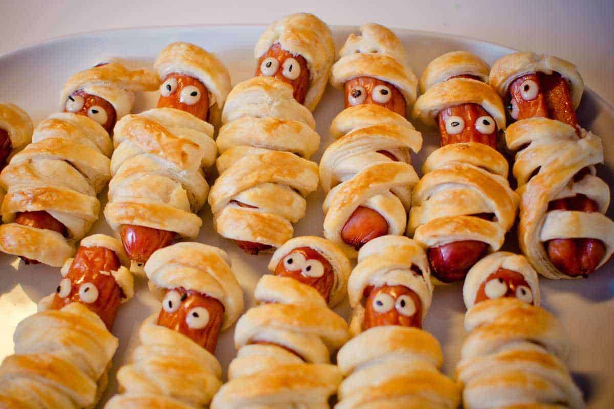 Bastoncini di salsiccia avvolti in pasta sfoglia con occhi di zucchero, preparati come finger food Halloween, presentati su un piatto bianco per una festa spettrale.