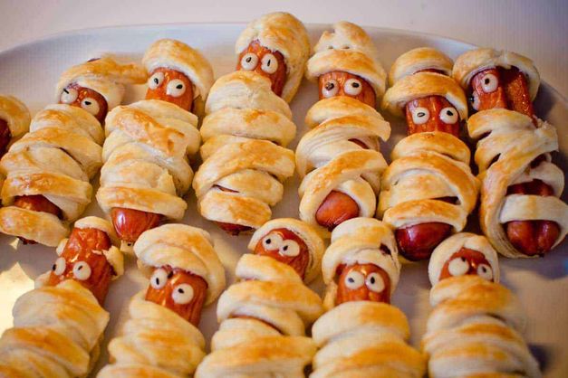 Finger foods: come preparare un aperitivo semplice e veloce 3 Bastoncini di salsiccia avvolti in pasta sfoglia con occhi di zucchero, preparati come finger food Halloween, presentati su un piatto bianco per una festa spettrale.