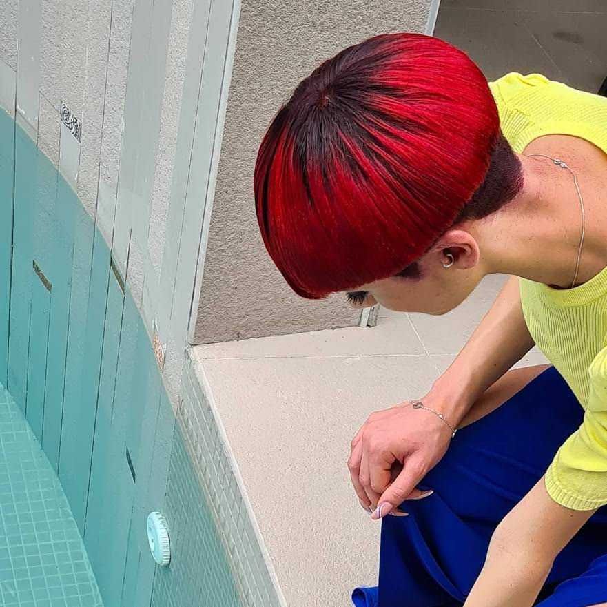 Colorata acconciatura bob rosso con sfumature nere, donna seduta vicino a piscina, stile moderno e audace, capelli lisci e lucidi, look casual con maglietta gialla e pantaloni blu, dettagli alla moda, immagini di lifestyle e bellezza, estetica di tendenza.