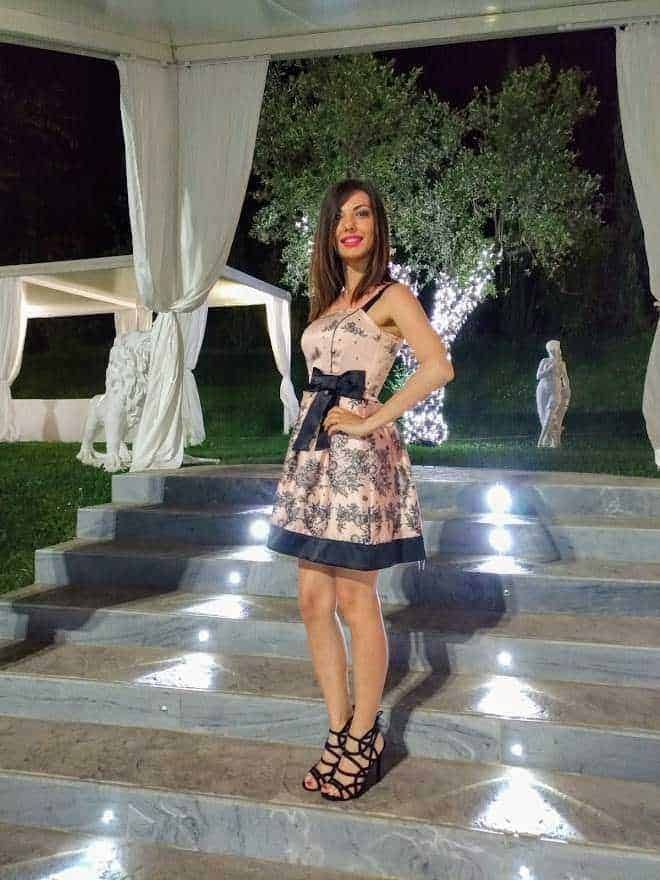 Elegante donna in abito blu in giardino con fontana, fiori e vegetazione, moda primavera estate, outfit femminile sofisticato, look da giorno in un parco fiorito, stile raffinato e chic.