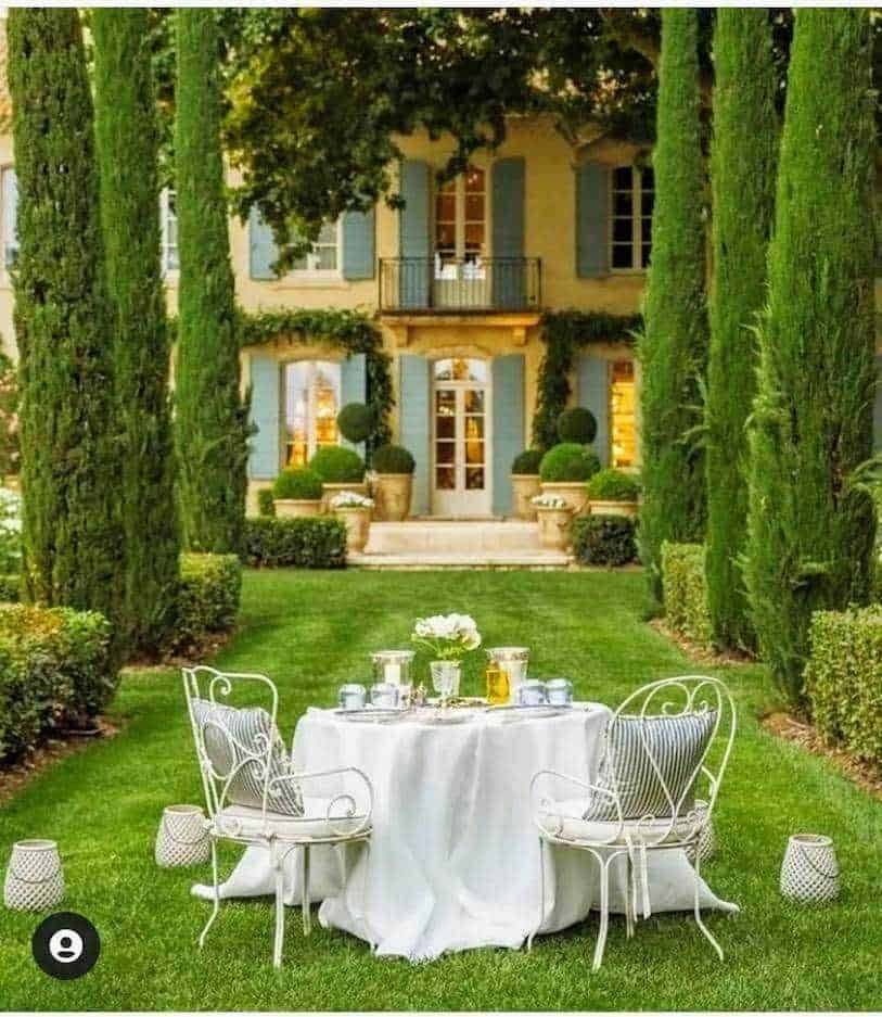 Elegante tavolo da colazione all'aperto in giardino, con sedie in ferro battuto, tovaglia bianca e decorazioni floreali, circondato da alberi e piante curate, perfetto per brunch estivi.