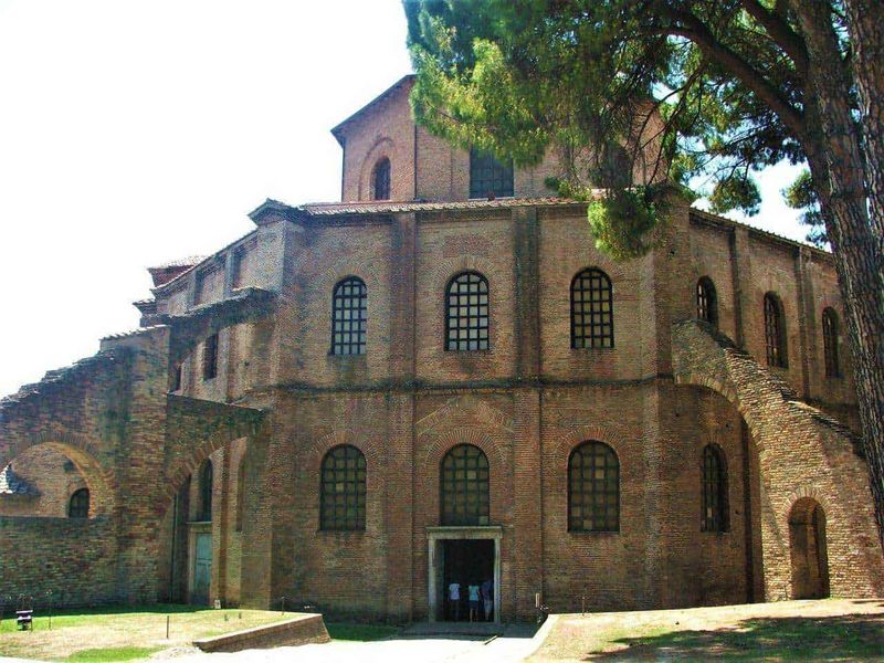 Basilica di San Vitale in Ravenna bizantina 2 Basilica di san vitale a ravenna: Antico edificio in stile romanico con mattoni a vista e finestre ad arco, circondato da alberi e paesaggio verde.