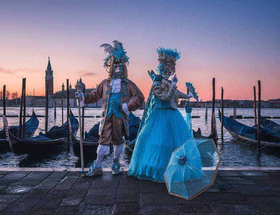 Maschere veneziane tradizionali con costumi eleganti e parasole lungo il canale a Venezia al tramonto. Scene tipiche del Carnevale di Venezia con gondole e skyline storico in sottofondo.