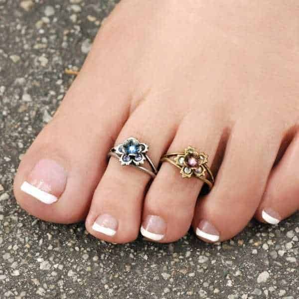 Storia dell'anello dei piedi significato e curiosità. Femminile piede con due anelli floreali in argento e ottone con pietre colorate, su sfondo di pietra. Perfetto esempio di gioielli alla moda e accessori estivi di tendenza. Bella decorazione per piede estivo.