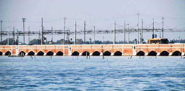Treno che attraversa un ponte sul mare con vista sull'acqua e linee di tralicci elettrici in background.