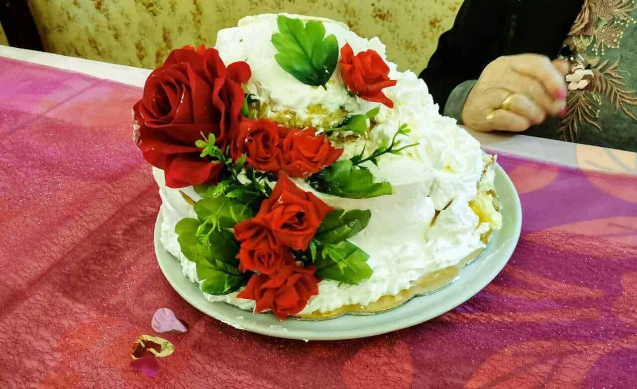 Torta decorata con roses rosse e foglie verdi, per occasioni speciali, presentata su un piatto bianco su tavolo con tovaglia rosa.