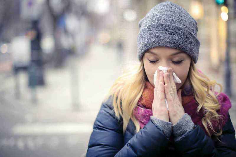 Ipotermia da COVID: 5 Sintomi Atipici e Quando Preoccuparsi (2026) 2 Naso di donna che si soffia il naso con fazzoletto in strada durante l'inverno, ambiente urbano freddo, malattia influenzale, donne con giacche e cappelli, influenza inverno.