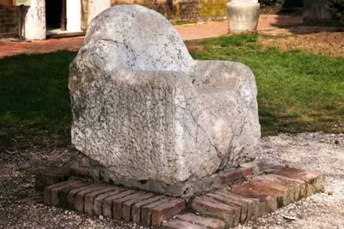 Sedia monumentale in pietra antica, decorativa e storica, posizionata su un basamento di mattoni, situata in un incantevole giardino con erba verde e piante.
