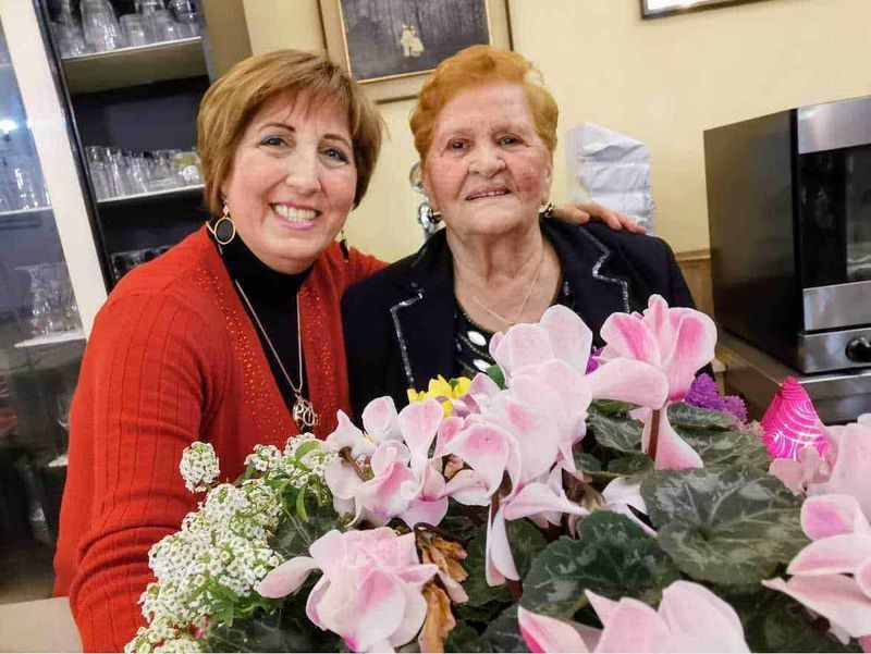 Quante rose si regalano per la festa della mamma? 3 Fiori di ciclamino rosa in primo piano con due donne anziane sorridenti sullo sfondo, ambientazione cucina o ristorante.