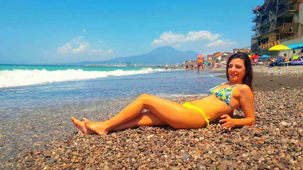 Spiaggia al mare con donna in bikini sdraiata sui ciottoli, ombrelloni colorati e cielo soleggiato, vista sul mare e sulle montagne sullo sfondo.