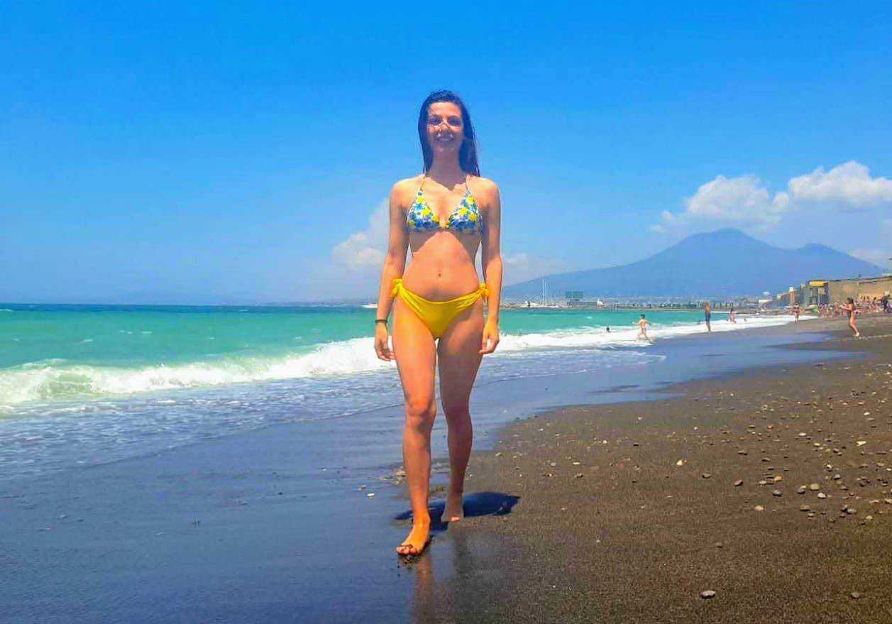 Ragazza in costume bikini sulla spiaggia al sole con mare e montagne sullo sfondo, perfetta scena di vacanza estiva e relax.