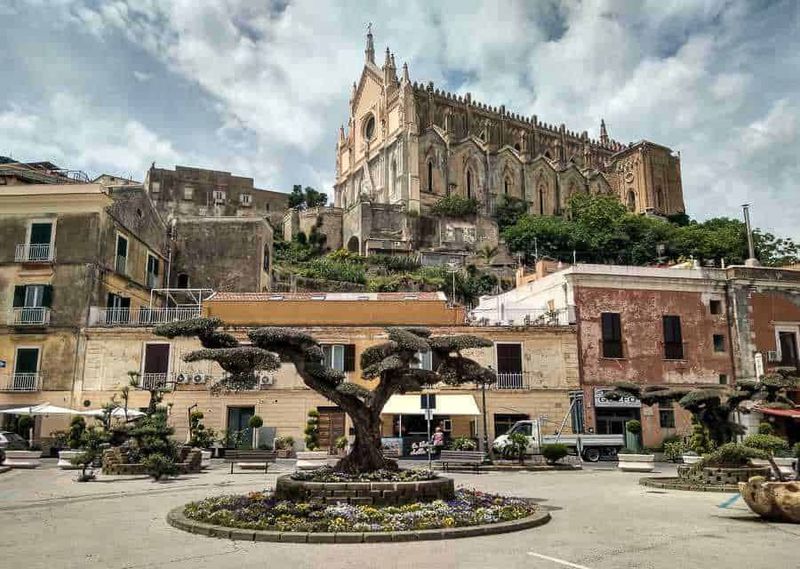 7 borghi tra Riviera di Ulisse, Monti Lepini e spiagge 15 Fontana centrale con alberi sagomati e fiori in primo piano, sullo sfondo una chiesa storica su una collina, suggestiva scena urbana con edifici colorati e cielo nuvoloso, immagine di suggestiva piazza italiana con monumento e architettura storica.