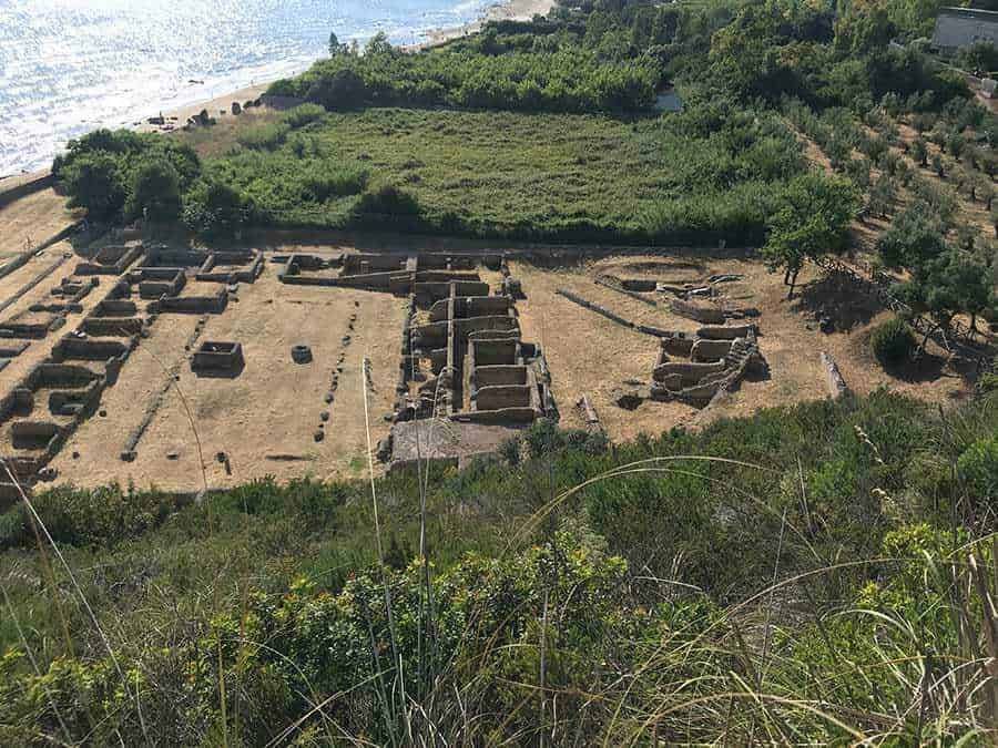Antica area archeologica con resti di mura e strutture in pietra, vista dalla collina, circondata da vegetazione e vicino al mare, sito di interesse storico e culturale in Italia, esploriamo le rovine storiche.
