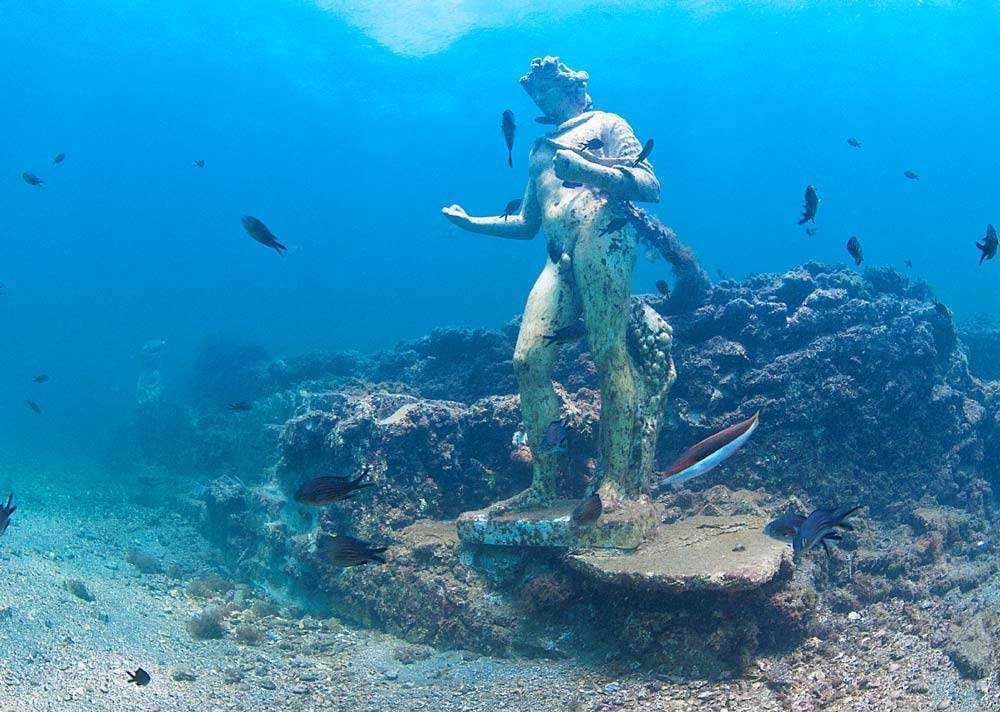 Scoprire una statua sommersa di una dea greca sotto il mare con pesci colorati e coralli, rappresentando l'arte e la storia subacquea. Perfetta per contenuti sull'esplorazione marina e il turismo sott'acqua.