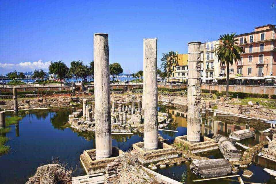 Baia sommersa a napoli: Resti archeologici di colonne romane vicino al mare, con edifici moderni sullo sfondo e un cielo sereno, rappresentano il patrimonio storico di Appunti Sul Blog.