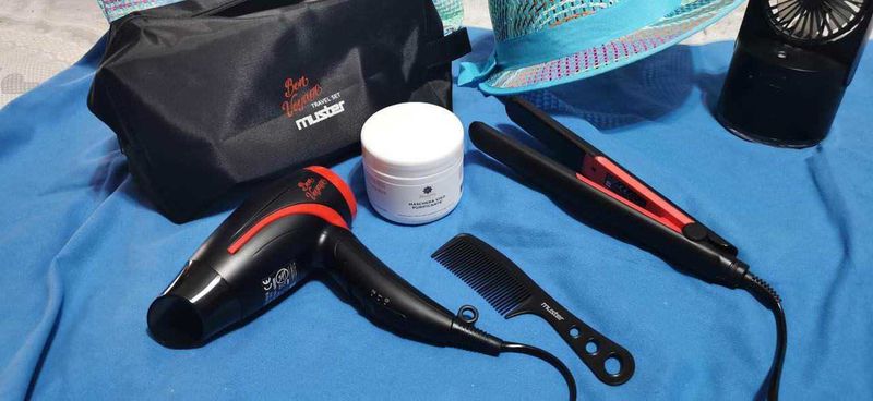 Cose da mettere in valigia utili in vacanza 2 Cosa mettere in valigia per la vacanza: Asciugacapelli nero e rosso con accessori per capelli, pettine e piastra per capelli, su asciugamano blu all'aperto, ready per cura e styling dei capelli.