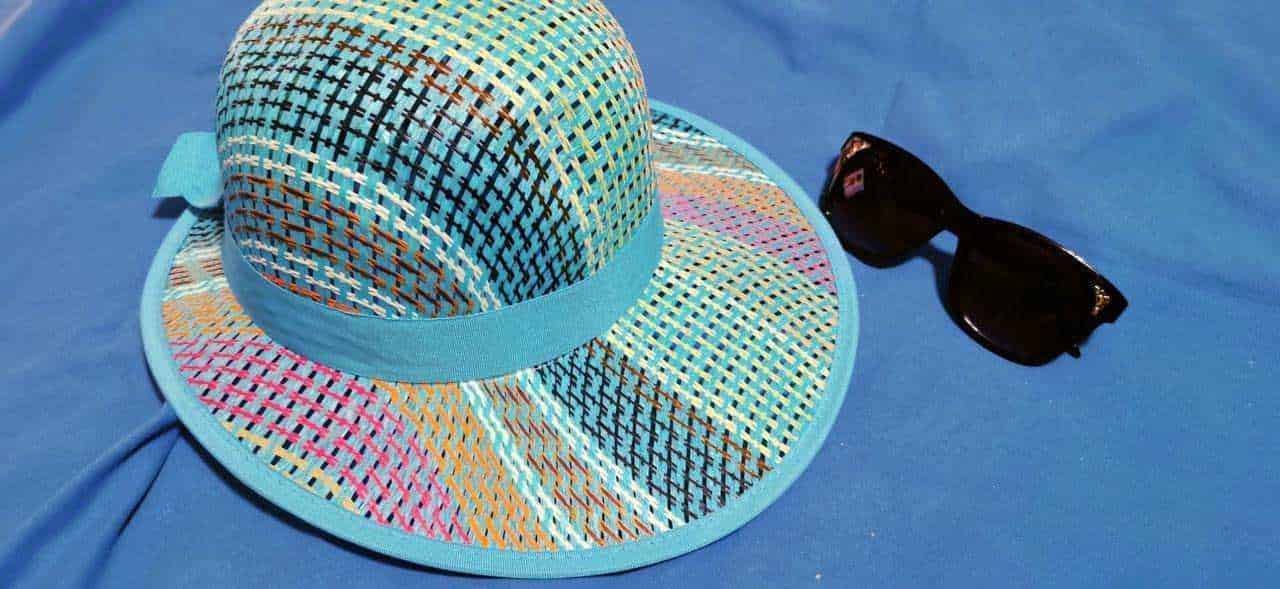 Cappello estivo di paglia colorato con nastro azzurro e occhiali da sole neri su sfondo blu.