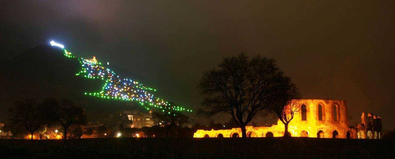 Colorato spettacolo di luci sul Monte Sacro con decorazioni natalizie e fuochi di artificio, evidenziando l'antico edificio in rovina e gli alberi circostanti durante la sera.
