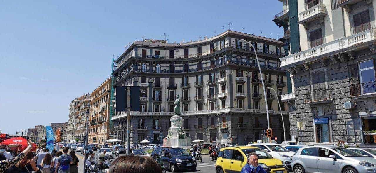 Automobilisti e pedoni su strada affollata di fronte a edifici storici in centro città, con traffico intenso e presenza di monumento in piazza, scena urbana vivace e trafficata.